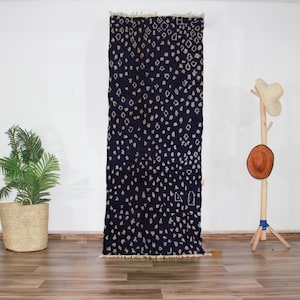 Peut inclure: Un tapis marocain bleu foncé avec un motif géométrique blanc. Le tapis a une bordure en franges et est suspendu à un mur blanc.