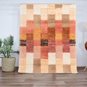 Op de afbeelding: Een kleurrijk, geometrisch patroon tapijt met vierkanten in tinten beige, bruin, oranje en roze. Het tapijt is gemaakt van pluche, textuur materiaal.