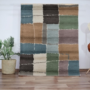 以下が含まれることがあります： 茶色、ベージュ、青、緑の正方形の幾何学模様が施された長方形の敷物。敷物は柔らかくふわふわした質感です。