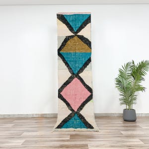 Puede incluir: Una alfombra larga y estrecha con un patrón geométrico en tonos de rosa, azul, dorado y negro sobre un fondo blanco.