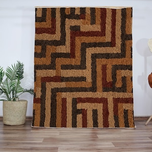Puede incluir: Una alfombra geométrica con patrones marrones, beige y rojos con una textura peluda. La alfombra tiene un diseño moderno con un patrón repetitivo de cuadrados y rectángulos.