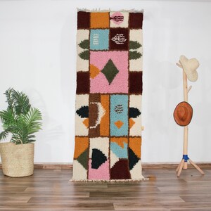 Peut inclure: Un tapis géométrique coloré avec un mélange de rose, bleu, marron et jaune. Le tapis est en laine et a une texture tissée à la main.