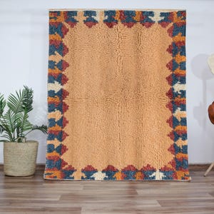 Authentic Moroccan Beni Ourain wool rug with warm ochre field and bold multicolor geometric border, handmade using 100% wool by Berber artisans; features rich earth tones of terracotta, blue, ivory, and rust; perfect for modern boho homes, Japandi, or rustic decor styles; this luxurious Moroccan wool carpet adds warmth and tribal charm to living rooms, dining rooms, or entryways while celebrating timeless craftsmanship from the Atlas Mountains.