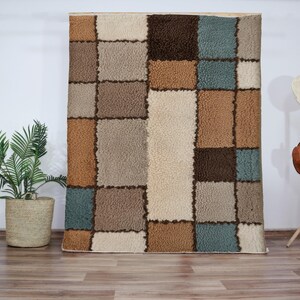 Peut inclure: Un tapis à motifs beige, marron et bleu avec une texture duveteuse. Le tapis est composé de carrés de différentes couleurs et tailles.