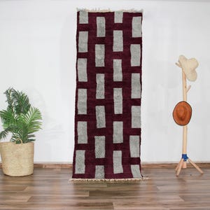 May include: A rectangular rug with a geometric pattern of gray rectangles on a burgundy background. The rug has a fringed edge and is displayed against a white wall. A potted plant and a wooden hat rack are also visible.