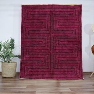 May include: A burgundy-coloured rectangular rug featuring a black geometric pattern. The rug is displayed against a white wall, with a potted plant and a hat stand in the background. The rug's design consists of connected, angular lines, creating a maze-like effect.