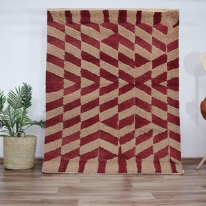 Op de afbeelding: Een rood en beige geometrisch tapijt met een chevron-patroon. Het tapijt is gemaakt van een zacht, pluche materiaal.