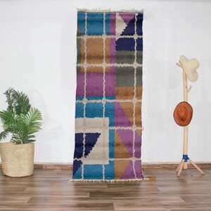 Peut inclure: Un tapis géométrique coloré avec des nuances de bleu, violet, marron et blanc. Le tapis a une texture tissée et est fait de laine.