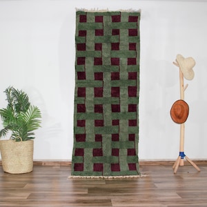 Peut inclure: Un tapis géométrique à motifs verts et bordeaux avec une texture tissée. Le tapis est suspendu à un mur blanc.