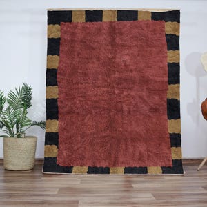 Peut inclure: Un tapis rouge avec une bordure géométrique noire et dorée. Le tapis a une texture douce et moelleuse.