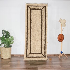May include: A long, rectangular rug with a white background and a brown border. The rug has a shaggy texture and is made of wool.