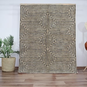 Op de afbeelding: Een beige en zwart geometrisch tapijt met een labyrint-achtig patroon. Het tapijt is gemaakt van een zacht, getextureerd materiaal en heeft een modern ontwerp.