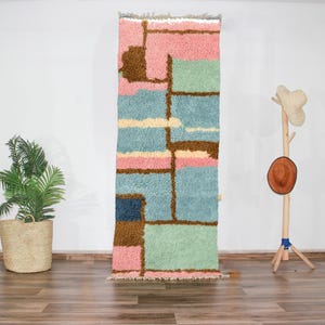 Peut inclure: Un tapis rectangulaire aux motifs géométriques. Il présente des blocs roses, bleus, verts et bruns. Le tapis est fait d'une matière douce et texturée. Il est présenté contre un mur blanc.