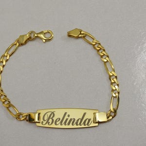 Puede incluir: Un brazalete de cadena dorada con una placa rectangular grabada con el nombre "Belinda".