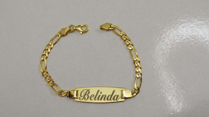 Puede incluir: Un brazalete de cadena dorada con una placa rectangular grabada con el nombre "Belinda".