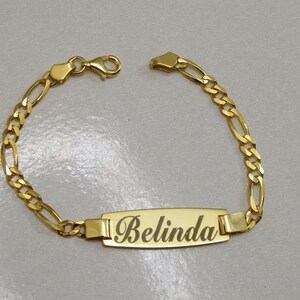 Puede incluir: Un brazalete de cadena dorada con una placa rectangular con el nombre "Belinda".