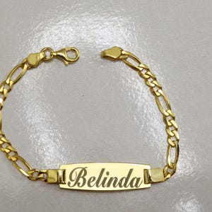 Puede incluir: Un brazalete de cadena dorada con una placa rectangular que dice "Belinda".