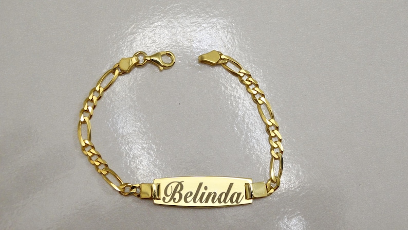Puede incluir: Un brazalete de cadena dorada con una placa rectangular que dice "Belinda".