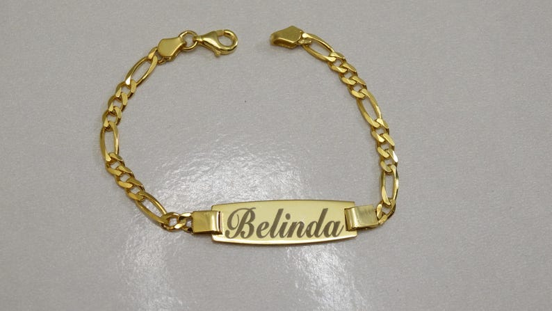 Puede incluir: Un brazalete de cadena dorada con una placa rectangular que dice "Belinda".