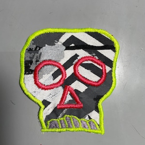 May include: A black and white skull patch with a geometric pattern. The skull has red eyes and a red triangle nose. The patch is outlined in neon green.