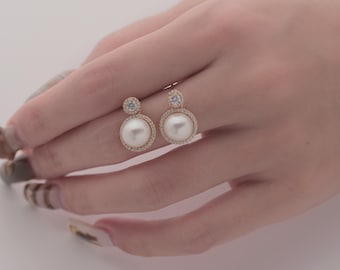 May include: A pair of gold earrings with a pearl and a small clear stone. The earrings are on a hand with manicured nails.