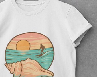 Puede incluir: Camiseta blanca con un diseño circular que representa una puesta de sol sobre las olas del océano y un surfista. Una concha marina está en primer plano. El texto "JUPITER FL" está debajo del diseño. La camiseta está colgada en una percha de madera.