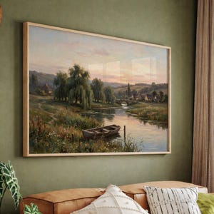 May include: A framed landscape painting depicts a serene river scene with a small boat, trees, and a bridge. The artwork features soft colors of green, brown, and blue, with a warm sunset sky. The frame is made of light-colored wood.