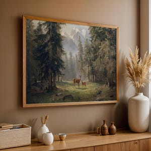 May include: A framed landscape painting depicts a forest scene with deer. The artwork, in a light wood frame, shows a mother deer and fawn in a clearing surrounded by tall trees and mountains in the background. The color palette is muted with greens, browns, and grays.