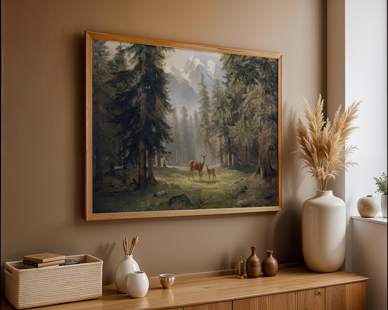 May include: A framed landscape painting depicts a forest scene with deer. The artwork, in a light wood frame, shows a mother deer and fawn in a clearing surrounded by tall trees and mountains in the background. The color palette is muted with greens, browns, and grays.
