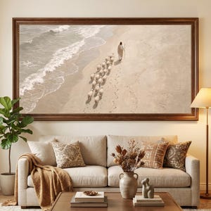 May include: A large rectangular framed wall art print depicting a figure in a light robe walking along a sandy beach shoreline followed by a small flock of sheep. The scene features soft beige and tan tones with white ocean waves crashing nearby.