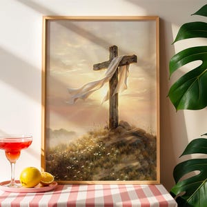 May include: A framed print depicts a weathered wooden cross draped with a white cloth, set against a sunset. The artwork is on a grassy hill with wildflowers. A cocktail and fruit sit on a pink and white checkered tablecloth.