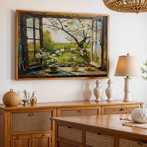 May include: A wooden framed painting depicts a scenic view through a window, showcasing a blossoming tree and a landscape. The painting hangs above a wooden cabinet with decorative items, including a lamp, vases, and a woven sphere. A woven light fixture hangs from the ceiling.