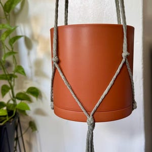 May include: Terracotta-colored ceramic plant pot suspended by a gray macrame hanger. The pot has a cylindrical shape and a small base. The macrame is knotted around the pot and has a tassel at the bottom. A green leafy plant is visible in the background.