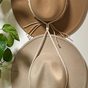 May include: Two brown felt hats hanging on a macrame wall hanger with a wooden ring at the top. The hats are different shades of brown and are hanging from the macrame cords.