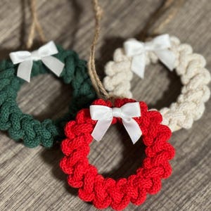 May include: Three small, knitted wreath ornaments in green, red, and cream colors, each adorned with a white bow. The wreaths are suspended by twine, suggesting they are for hanging as holiday decorations.