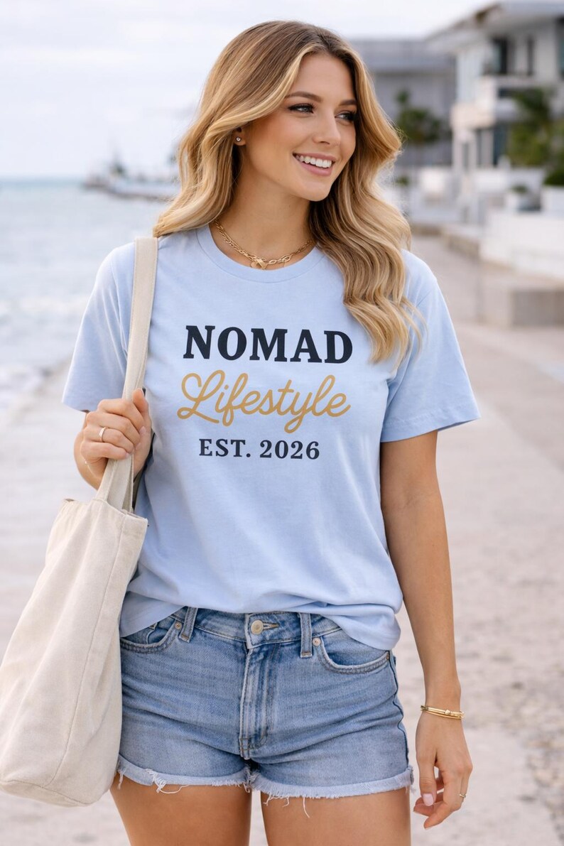 Blonde woman smiling on a waterfront promenade wearing a light blue “NOMAD Lifestyle EST. 2026” graphic T-shirt, denim shorts, and gold jewellery, holding a beige tote bag with water and modern buildings in the background.