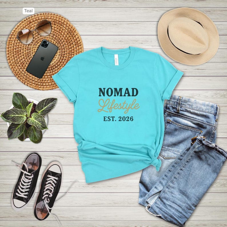 May include: Teal crew neck t-shirt with the words "NOMAD Lifestyle EST. 2026" in black and gold. The shirt is paired with blue jeans, black trainers, a straw hat, sunglasses, and a smartphone on a woven mat.