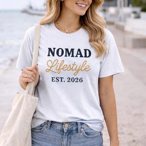 Blonde woman smiling on a waterfront promenade wearing a white “NOMAD Lifestyle EST. 2026” graphic T-shirt, denim shorts, and gold jewellery, holding a beige tote bag with water and modern buildings in the background.