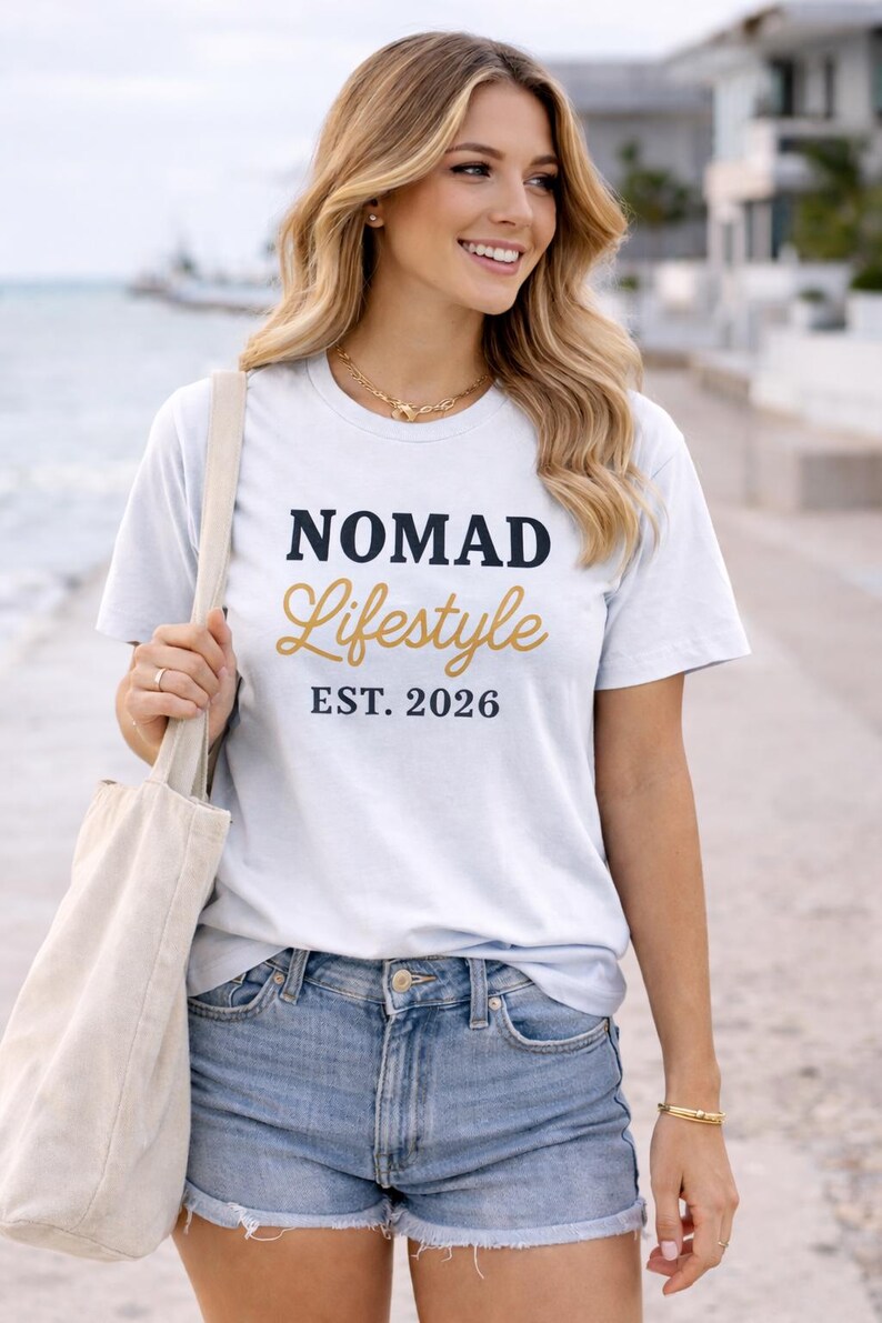 Blonde woman smiling on a waterfront promenade wearing a white “NOMAD Lifestyle EST. 2026” graphic T-shirt, denim shorts, and gold jewellery, holding a beige tote bag with water and modern buildings in the background.