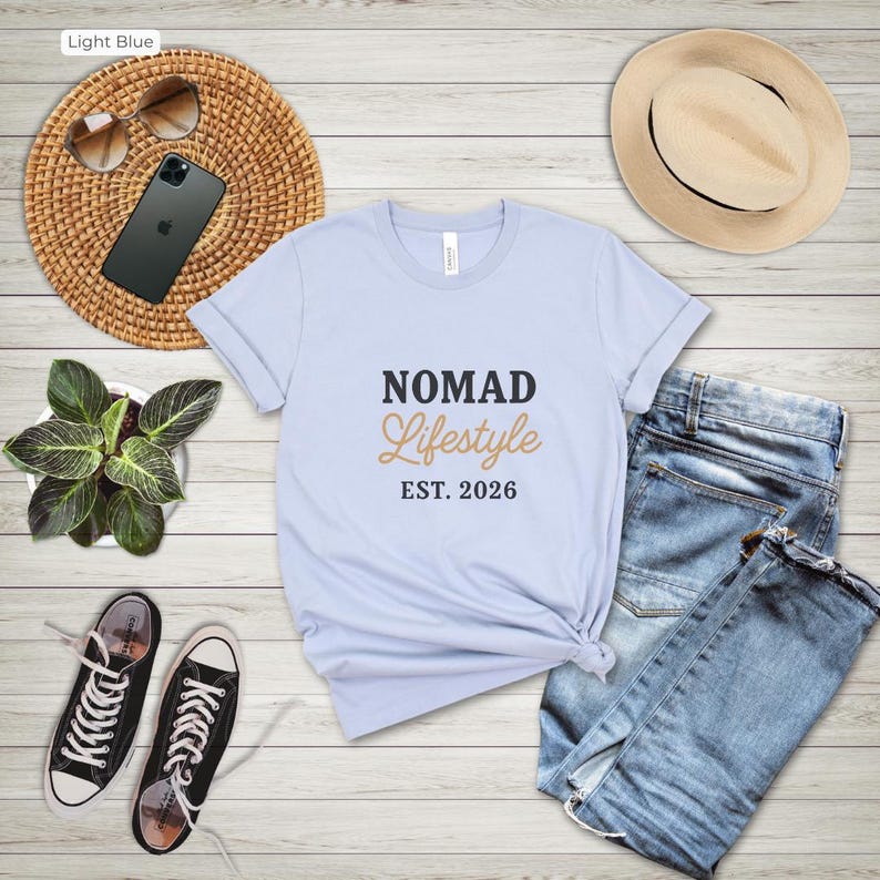 May include: A pale blue t-shirt with the text "NOMAD LIFESTYLE EST. 2026". The shirt is styled with blue denim jeans, black trainers, a straw hat, and sunglasses. A smartphone and a small potted plant are also visible.