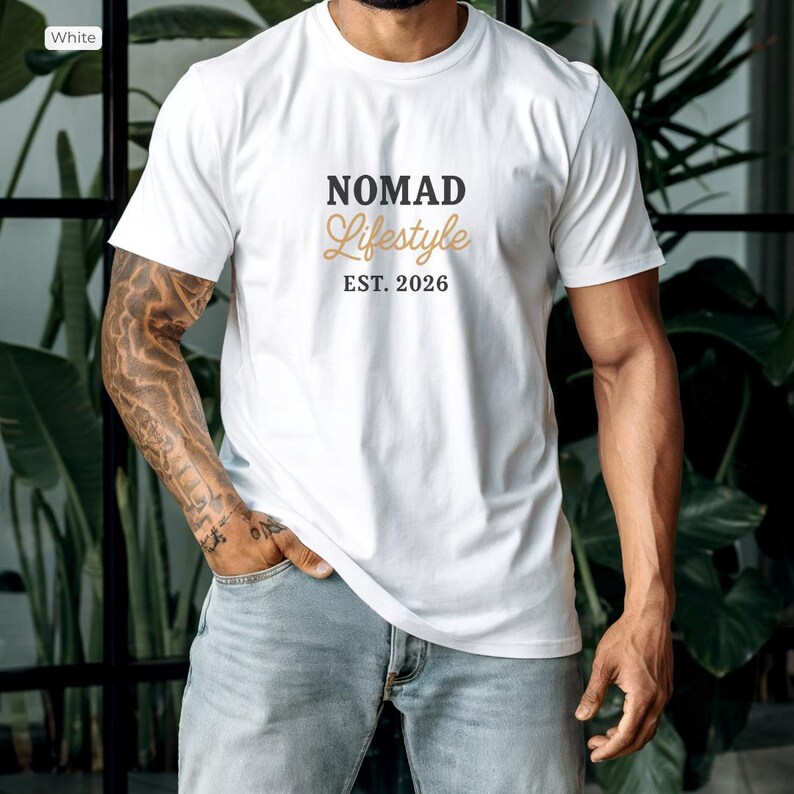 May include: White crew neck t-shirt featuring the text "NOMAD Lifestyle EST. 2026" in black and gold. The shirt is worn with light blue jeans. The image is set against a backdrop of greenery.