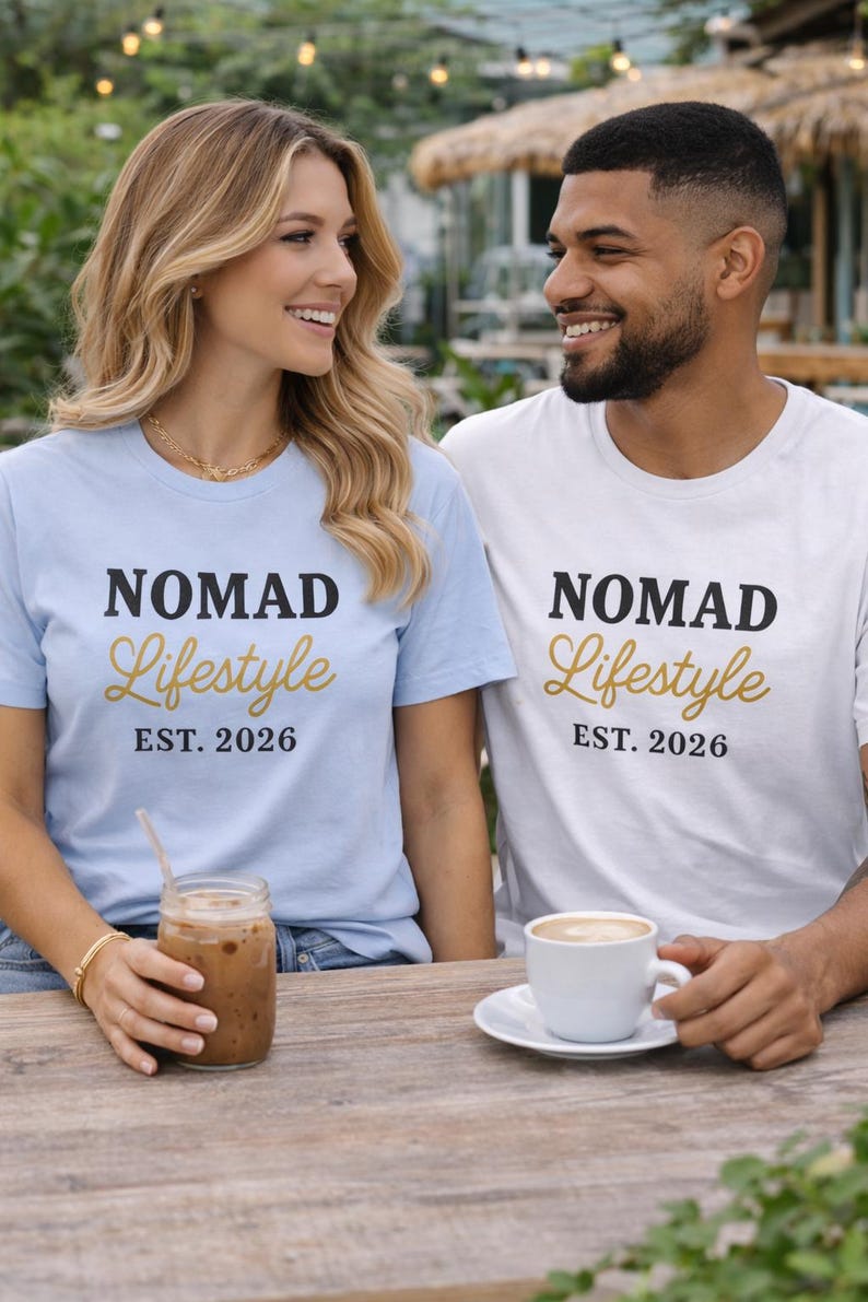 Man and woman smiling on a waterfront promenade wearing a white and a light blue “NOMAD Lifestyle EST. 2026” graphic T-shirt, denim shorts, and gold jewellery, holding drinks on a table.