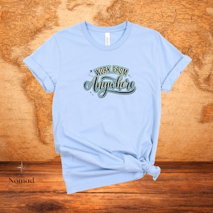 Pu&ograve; includere: T-shirt azzurra con maniche arrotolate. La maglietta presenta la scritta "WORK FROM Anywhere" in un design verde e bianco. La maglietta &egrave; annodata in basso. Lo sfondo &egrave; una mappa del mondo.