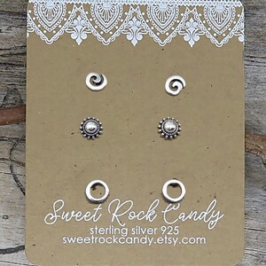 May include: Three pairs of sterling silver 925 stud earrings displayed on a rectangular tan card with white lace-style trim. The earrings feature spiral, sunburst, and open circle designs. The card includes the text Sweet Rock Candy and website.