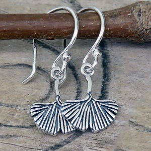 May include: Silver-toned earrings featuring a ginkgo leaf design. The earrings are suspended from a hook and have a detailed, fan-shaped leaf with intricate veining. The earrings are displayed against a wooden background.