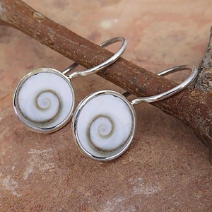 May include: A pair of round earrings featuring white shiva eye shells with a natural brown spiral pattern, set in polished silver-toned metal frames with curved ear wires. The circular shell centers are smooth and display a distinct, centered swirl.