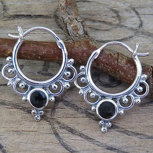 May include: A pair of silver hoop earrings featuring a circular black stone at the base, surrounded by intricate metal scrollwork and small silver beads. The earrings have a hinged closure mechanism and a polished finish on the metal surface.
