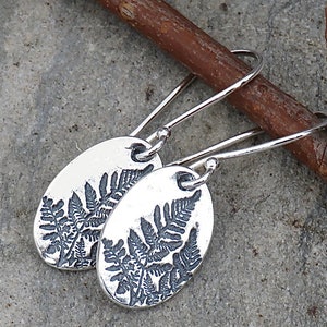 May include: A pair of silver earrings with a fern design. The earrings are oval shaped and have a hook closure.