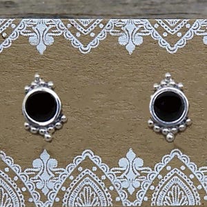 May include: A pair of round sterling silver stud earrings with black centers, featuring a decorative beaded border. The earrings are attached to a rectangular brown display card with white lace-style borders and the text Sweet Rock Candy.