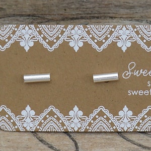 May include: Two small, cylindrical sterling silver stud earrings attached to a rectangular brown cardboard display card. The card features a white lace-like border pattern and white text that reads Sweet Rock Candy, sterling silver .925, and sweetrockcandy.etsy.com.